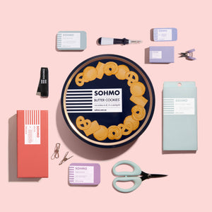 Collection of SOHMO branded packaging and cookie tins on a pink background