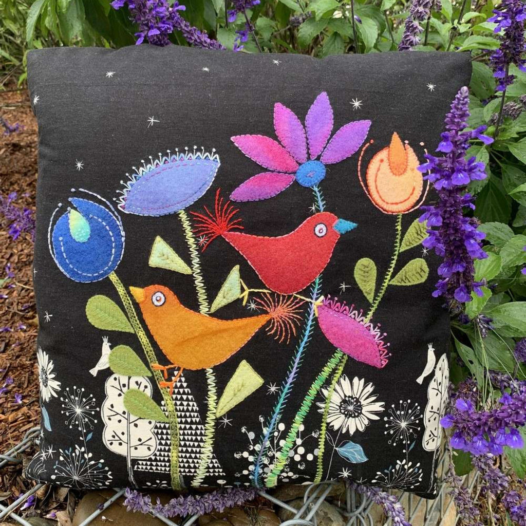 Decorative pillow with colorful birds and flowers on a black background, surrounded by purple flowers.