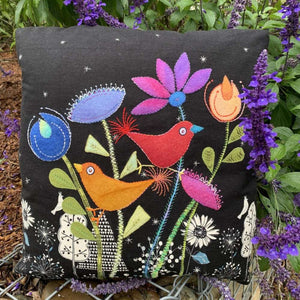 Decorative pillow with colorful birds and flowers on a black background, surrounded by purple flowers.